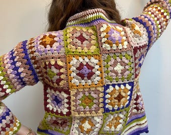 Puede incluir: Una chaqueta de punto de ganchillo colorida con un patrón de cuadrados de la abuela en tonos de morado, verde, marrón y crema. La chaqueta tiene cuello alto y mangas largas, y está acabada con un ribete morado.