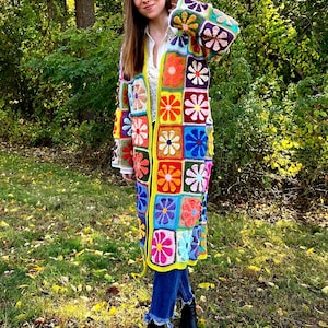 May include: A long, colorful crochet cardigan featuring square floral patterns in various colors. The cardigan has a yellow border and is worn over a white shirt and blue jeans. The overall style is bohemian and handmade.