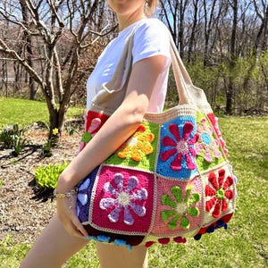 May include: A large, colorful crochet tote bag with a patchwork design. The bag features squares of various colors, each adorned with a floral motif. The bag has a beige strap and trim, and is held by a person.
