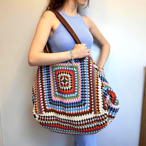 May include: A large, slouchy tote bag crafted from multicolored crochet yarn in a square granny pattern. The bag features a wide, brown leather shoulder strap and a vibrant design incorporating shades of blue, red, orange, pink, and cream.