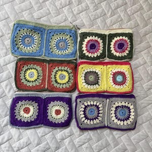 May include: Six colorful crochet pouches with zipper closures are displayed. Each pouch features a square design with a central floral motif. The color palette includes blue, green, red, yellow, purple, and gray, offering a variety of options.