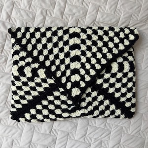 May include: A black and cream-colored crocheted clutch with a geometric pattern. The handmade bag features a flap closure and a checkerboard design. The crochet stitches create a textured surface, perfect for carrying essentials.