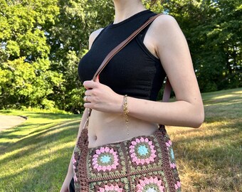May include: A brown, pink, green, and blue crocheted shoulder bag. The bag features a floral pattern with square motifs. The strap is brown leather. The bag is worn over the shoulder.