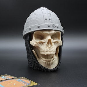 May include: A skull figurine wearing a gray helmet and chainmail. The skull is off-white with visible teeth and eye sockets. The helmet has rivets and a rounded shape. The chainmail is black. A few trading cards are in the foreground.