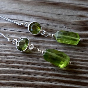 May include: A pair of silver earrings featuring a round, faceted peridot stone and a rectangular, faceted peridot stone. The earrings are on a silver chain and are set against a rustic wooden background.