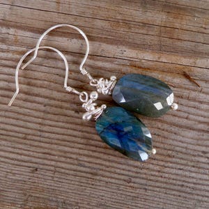 May include: A pair of silver-toned earrings with faceted, oval-shaped blue-green gemstones. The earrings have a simple hook design and small decorative silver accents near the gemstones. The gemstones have a polished, reflective surface.