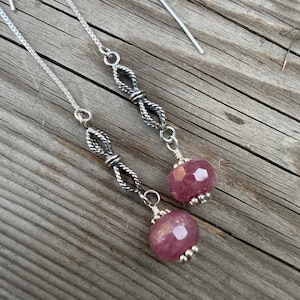 May include: A pair of silver threader earrings featuring a delicate chain, a twisted metal infinity-style link, and a faceted, round, deep pink gemstone bead at the base. The earrings are constructed with a long, thin silver wire for threading.