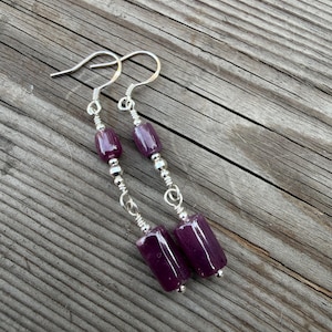 May include: A pair of drop earrings featuring silver-toned hooks and wire-wrapped connections. Each earring consists of a small cylindrical dark purple bead, a tiny silver-toned spacer bead, and a larger cylindrical dark purple bead at the base.