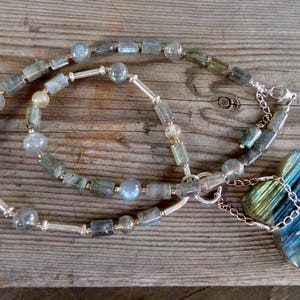 May include: A handcrafted necklace featuring a mix of blue, green, and gray labradorite beads. The necklace includes a large, cross-shaped pendant with a silver chain and clasp. The beads are strung on a silver chain, creating a unique jewelry piece.