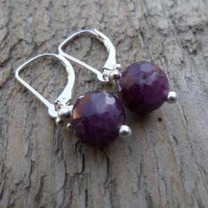 May include: A pair of silver earrings with purple gemstone beads. The earrings have a simple design with a small silver loop at the top and a larger silver loop at the bottom.
