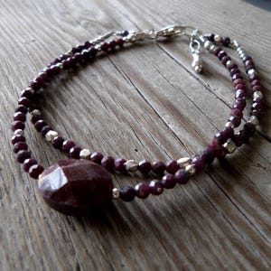 May include: A double-strand bracelet featuring faceted, deep red beads and small silver-colored beads. A large, faceted, oval-shaped, deep red stone is centered on the bracelet. The bracelet is displayed on a wooden surface.