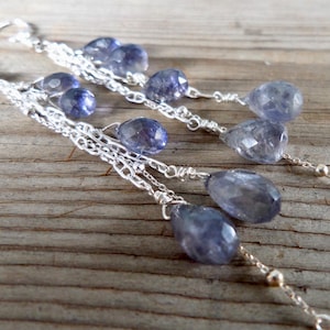 May include: Silver chain earrings with multiple strands, each adorned with faceted, teardrop-shaped, blue-violet gemstones. The earrings are displayed on a rustic wooden surface, showcasing their delicate design and color.