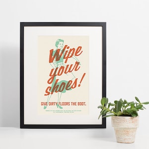 Może przedstawiać: Oprawiony plakat w stylu vintage z napisem "Wipe your shoes!" w czerwonym skrypcie na turkusowej ilustracji kobiety. Poniżej znajduje się fraza "GIVE DIRTY FLOORS THE BOOT". Plakat jest w czarnej ramie.