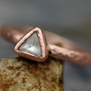 May include: A close-up of a copper ring with a triangular, clear gemstone. The ring has a hammered texture on the band and a bezel setting around the stone. The ring is set on a small, textured rock.