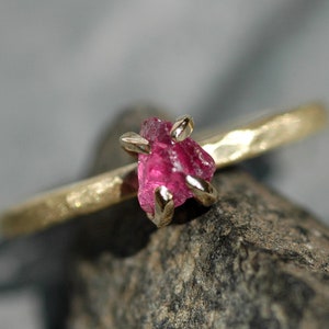 May include: A gold ring with a raw pink ruby gemstone set in the centre. The ring has a hammered texture.
