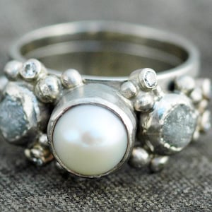 May include: A silver ring with a large white pearl and smaller white gemstones. The ring has a textured, organic design.