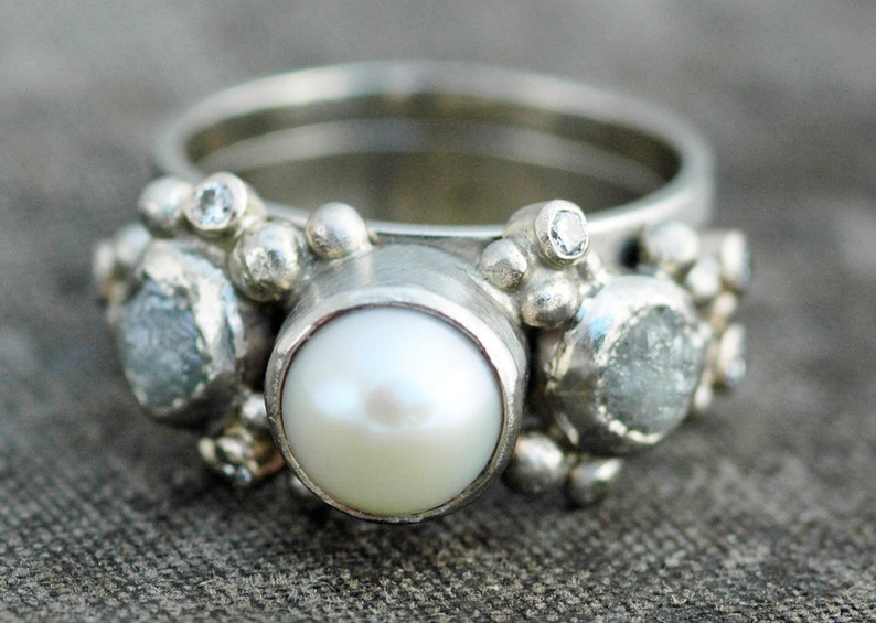 May include: A silver ring with a large white pearl and smaller white gemstones. The ring has a textured, organic design.