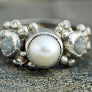May include: A silver ring with a large white pearl in the center and smaller diamonds on either side. The ring is set with a cluster of small silver beads.