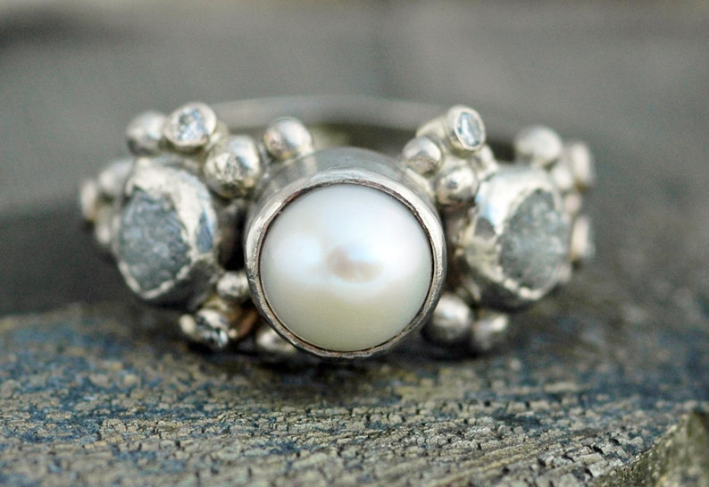 May include: A silver ring with a large white pearl in the center and smaller diamonds on either side. The ring is set with a cluster of small silver beads.