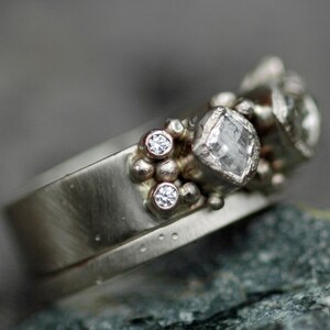 May include: A silver ring with a cluster of small diamonds and a larger, square-cut diamond. The ring is set on a textured band.