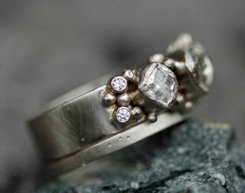 May include: A silver ring with a cluster of small diamonds and a larger, square-cut diamond. The ring is set on a textured band.