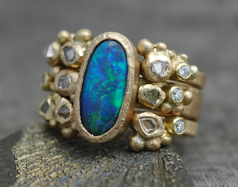 K&ouml;nnte beinhalten: Ein goldener Ring mit einem gro&szlig;en ovalen Opal-Edelstein, umgeben von kleineren Diamanten. Der Ring besteht aus mehreren B&auml;ndern mit strukturierten Golddetails.