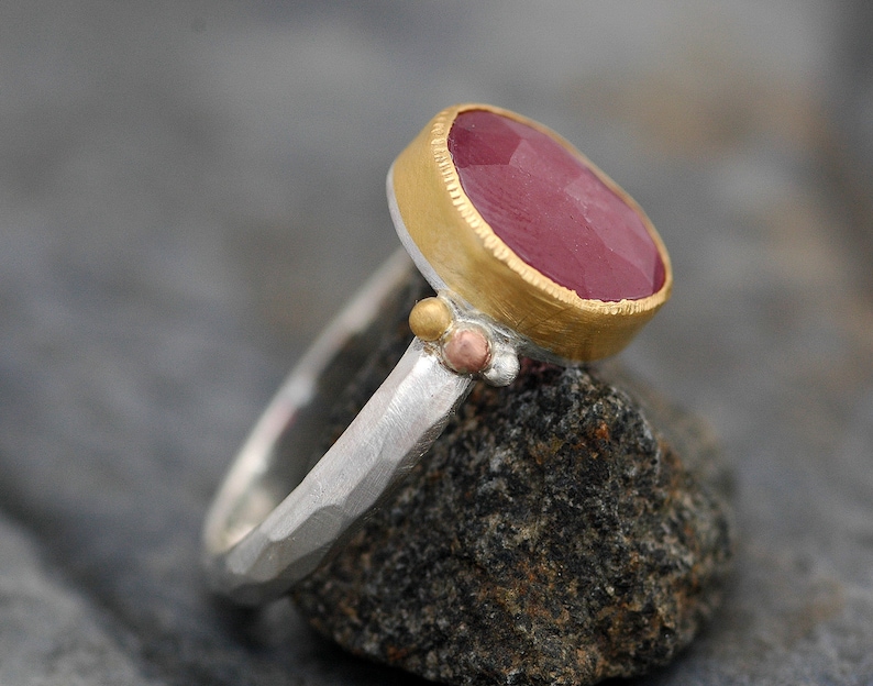 May include: A silver ring with a hammered texture and a pink gemstone set in a gold bezel. The ring is accented with small gold and copper beads.