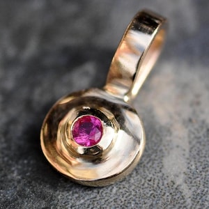 May include: A gold pendant with a round, textured surface and a small pink gemstone in the center.