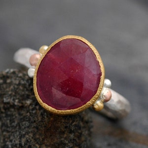 May include: A ring with a large, oval-shaped ruby gemstone set in a gold bezel. The ring band is made of silver and has three small gold beads on either side of the stone.