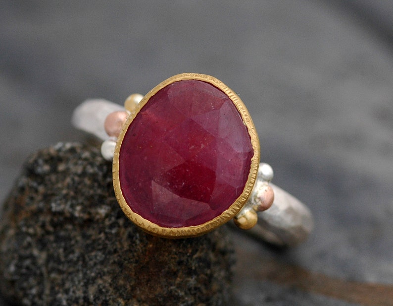 May include: A ring with a large, oval-shaped ruby gemstone set in a gold bezel. The ring band is made of silver and has three small gold beads on either side of the stone.