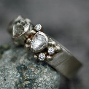 May include: A silver ring with a cluster of small diamonds and two larger, rough-cut diamonds. The ring is set on a textured band.