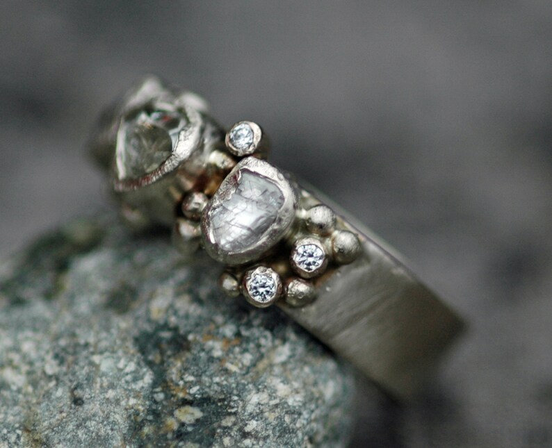 May include: A silver ring with a cluster of small diamonds and two larger, rough-cut diamonds. The ring is set on a textured band.