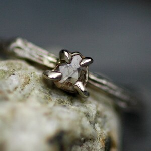 May include: A silver ring with a rough, clear diamond set in a prong setting. The ring is on a grey, textured surface.