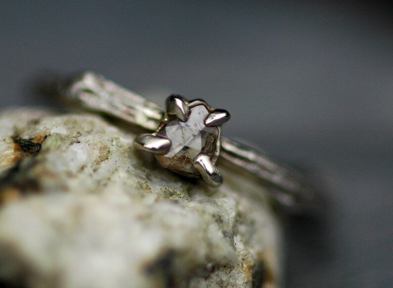 May include: A silver ring with a rough, clear diamond set in a prong setting. The ring is on a grey, textured surface.