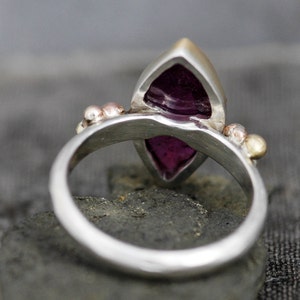 May include: A silver ring with a purple gemstone set in a marquise shape. The ring has small gold and silver accents.