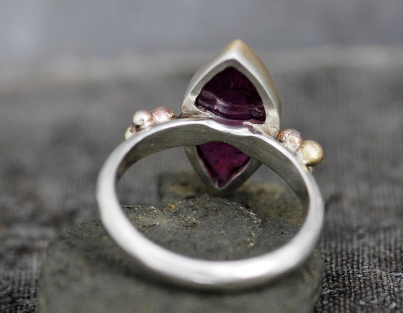 May include: A silver ring with a purple gemstone set in a marquise shape. The ring has small gold and silver accents.
