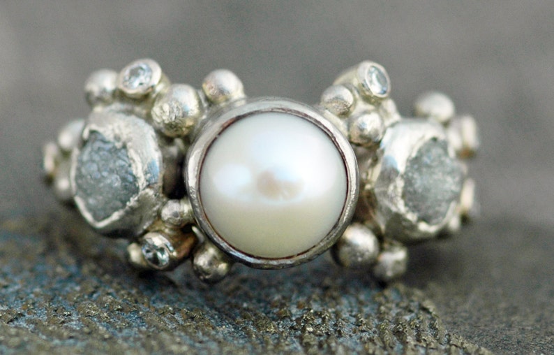 May include: A silver ring with a large white pearl in the center, surrounded by smaller white stones and silver beads.