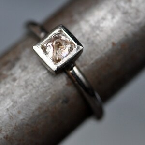 May include: A silver ring with a square-shaped, clear, faceted gemstone set in a silver bezel.