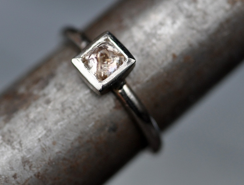May include: A silver ring with a square-shaped, clear, faceted gemstone set in a silver bezel.