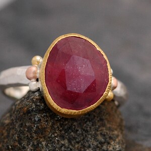 May include: A silver ring with a large, faceted ruby gemstone set in a gold bezel. The ring is decorated with small gold and copper beads.