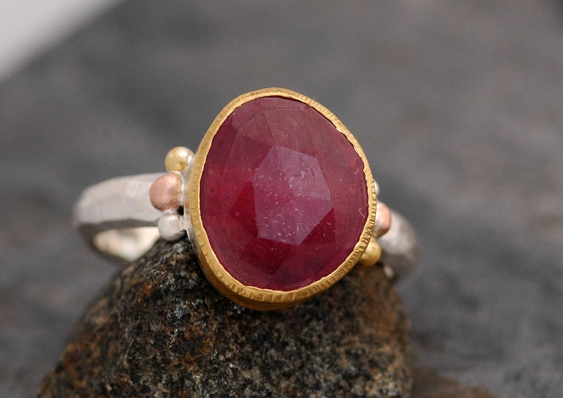 May include: A silver ring with a large, faceted ruby gemstone set in a gold bezel. The ring is decorated with small gold and copper beads.