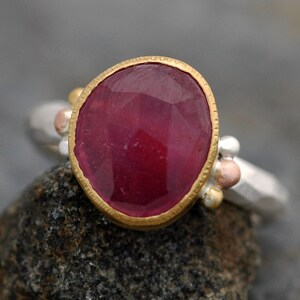 May include: A ring with a large, faceted ruby gemstone set in a gold bezel. The ring band is made of silver and has small gold and copper beads.