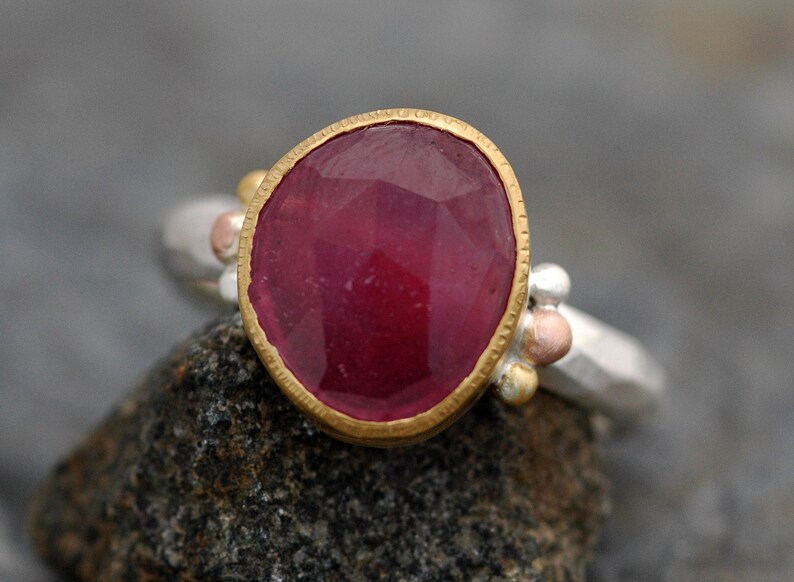 May include: A ring with a large, faceted ruby gemstone set in a gold bezel. The ring band is made of silver and has small gold and copper beads.