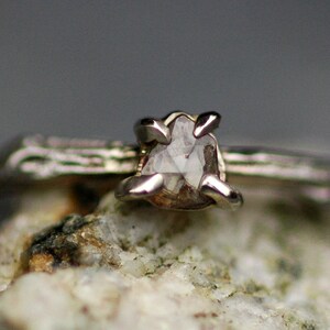 May include: A silver ring featuring a rough, clear diamond set in a prong setting. The ring is resting on a piece of rough, white rock.