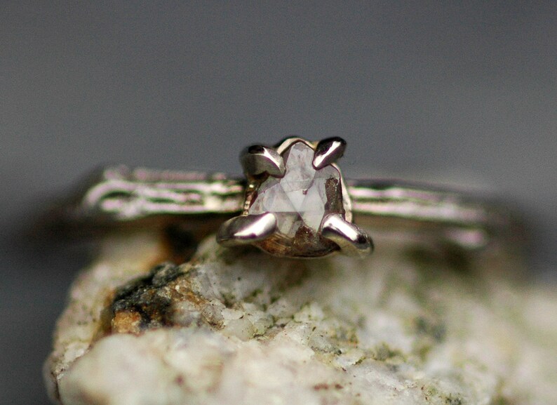 May include: A silver ring featuring a rough, clear diamond set in a prong setting. The ring is resting on a piece of rough, white rock.