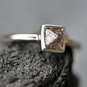May include: A silver ring with a square setting featuring a rough, clear diamond.