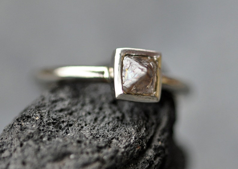 May include: A silver ring with a square setting featuring a rough, clear diamond.