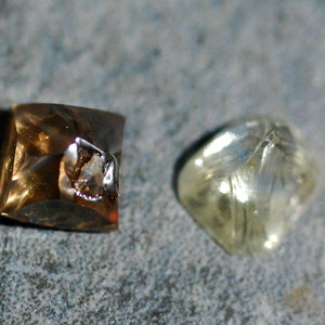 May include: Three faceted gemstones, one brown, one clear, and one yellow, are displayed on a gray surface.