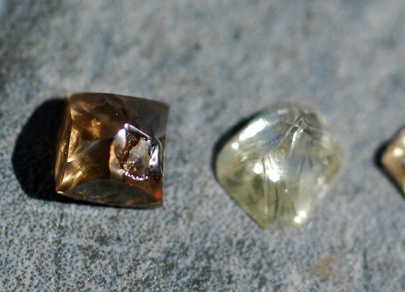 May include: Three faceted gemstones, one brown, one clear, and one yellow, are displayed on a gray surface.