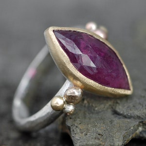 May include: A silver ring with a marquise-shaped ruby gemstone set in a gold bezel. The ring is accented with small gold beads.